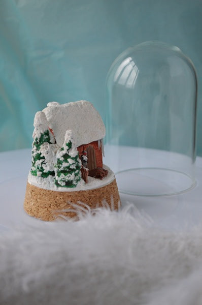 Miniature snowy scene with a cabin under a glass dome on a white surface.