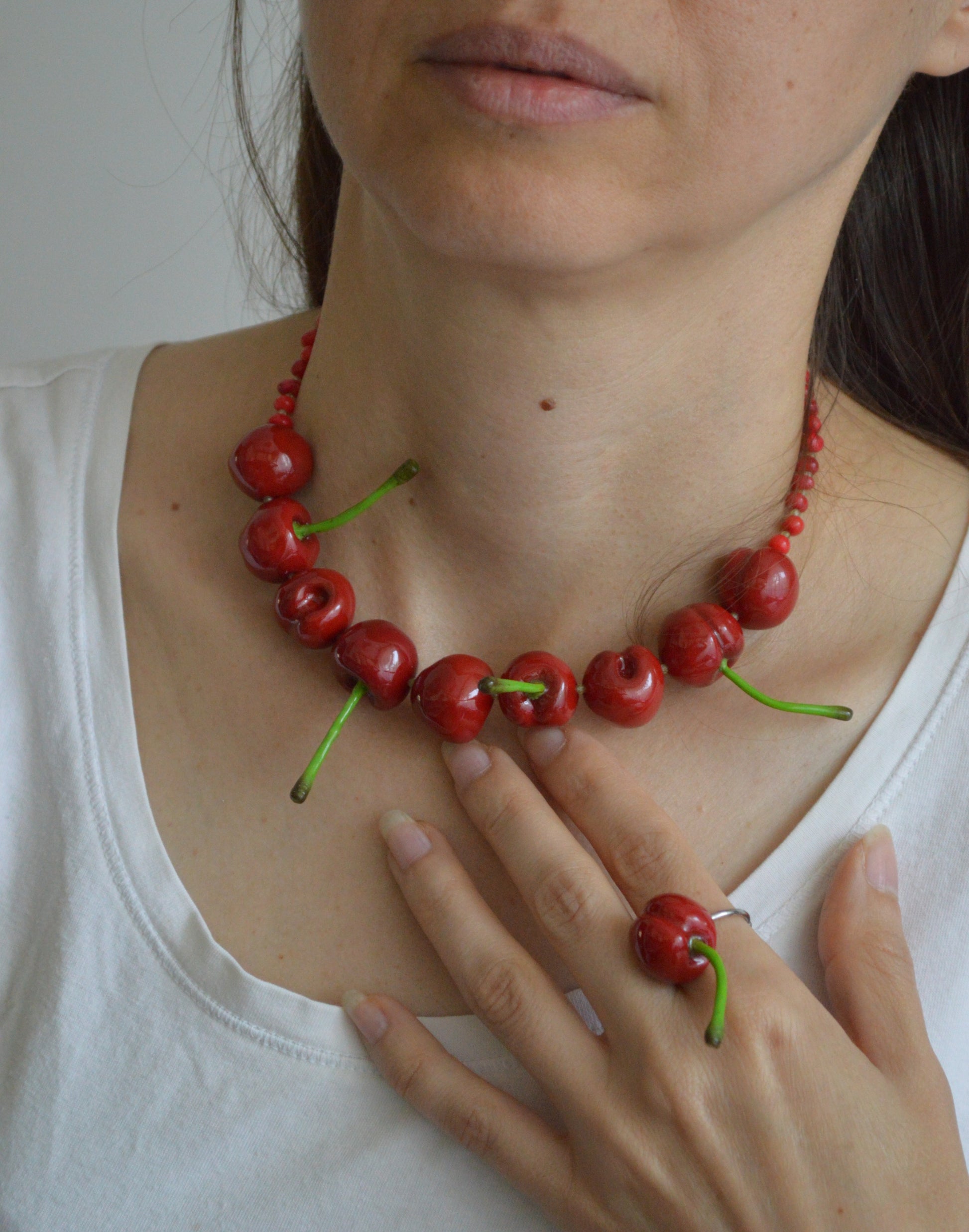 Person wearing a necklace and ring made of red cherries with green stems on a plain background