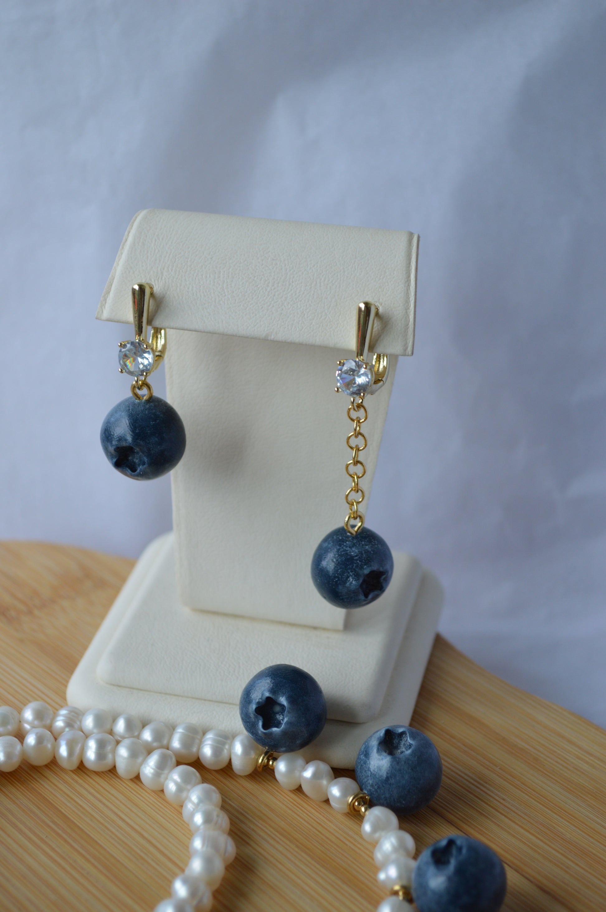 Blueberry-themed earrings on a white stand with a pearl necklace in the foreground.