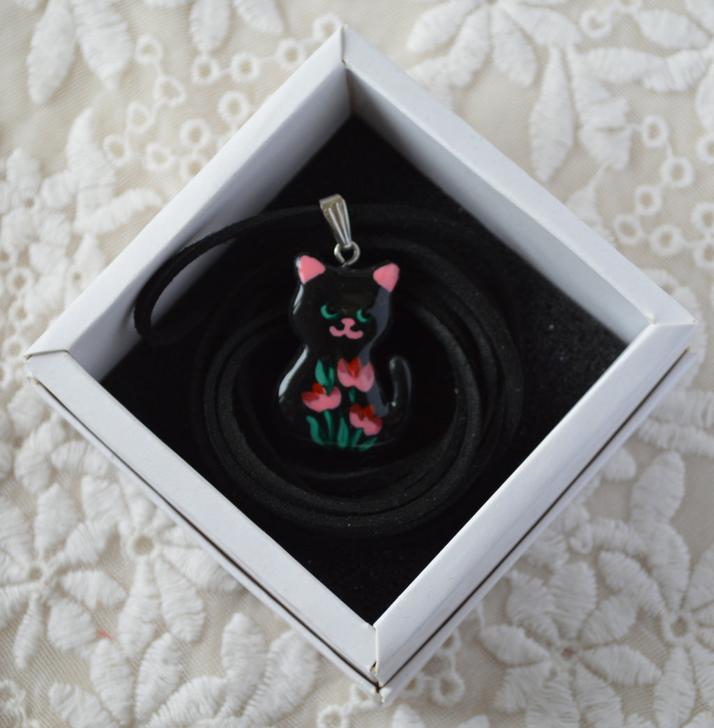 Black cat-shaped pendant with pink accents in a white box on a lace background