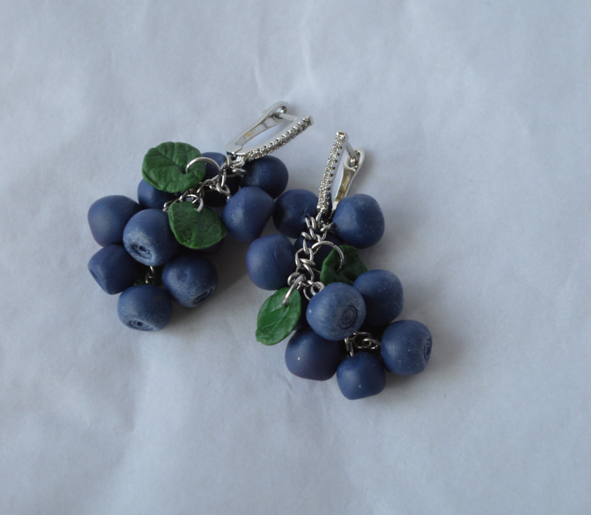 Blue berry-shaped earrings with green leaves on a light gray background